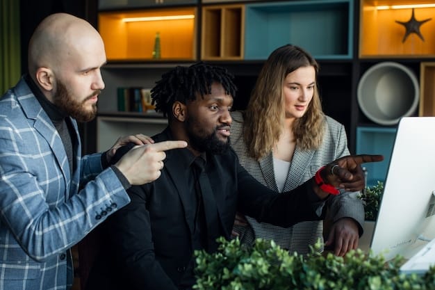 A group of diverse people sitting around a table in a modern office space, collaborating on a project. Laptops and tablets are visible on the table, suggesting a tech-driven environment.