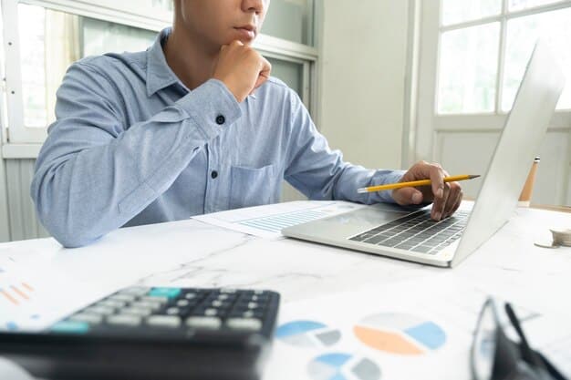 A person using a calculator to plan finances with various documents and a laptop displaying financial charts on a desk. Focus is on the focused expression of the individual.